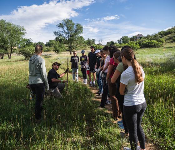 Group of college students standing on a trail in a grassy meadow listen and watch a demonstration.. 